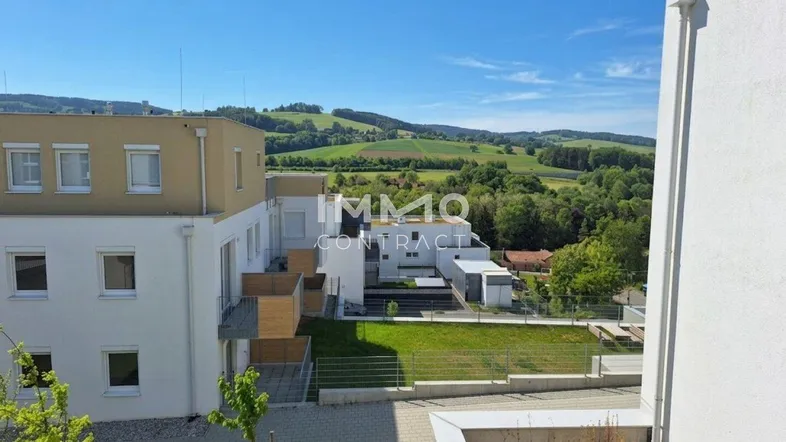 Blick von einem Balkon auf moderne Wohngebäude und eine grüne Hügellandschaft unter blauem Himmel.