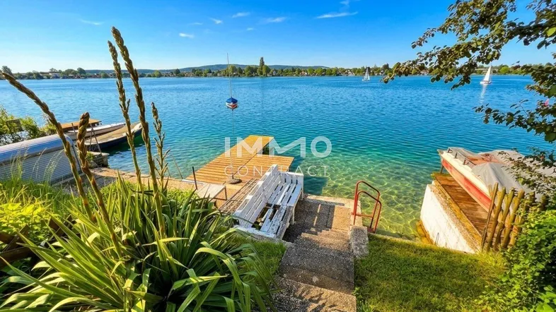 Malerischer Blick auf den See mit Steg, Booten und üppiger Ufervegetation unter blauem Himmel.