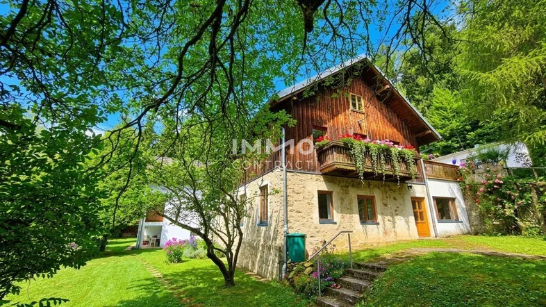Traditionelles Landhaus mit Holzbalkon und Steinfassade, umgeben von üppigem Grün und Bäumen.
