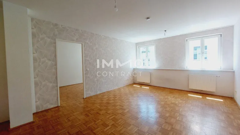 Geräumiges Wohnzimmer mit Parkettboden, gemusterter Tapete und zwei großen Fenstern.
