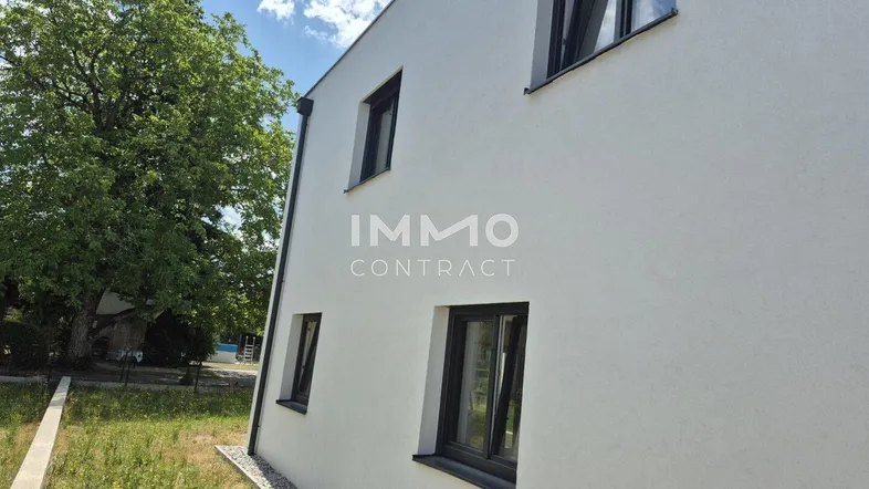 Moderne Hausfassade mit weißen Wänden und dunklen Fensterrahmen, umgeben von Grünflächen.