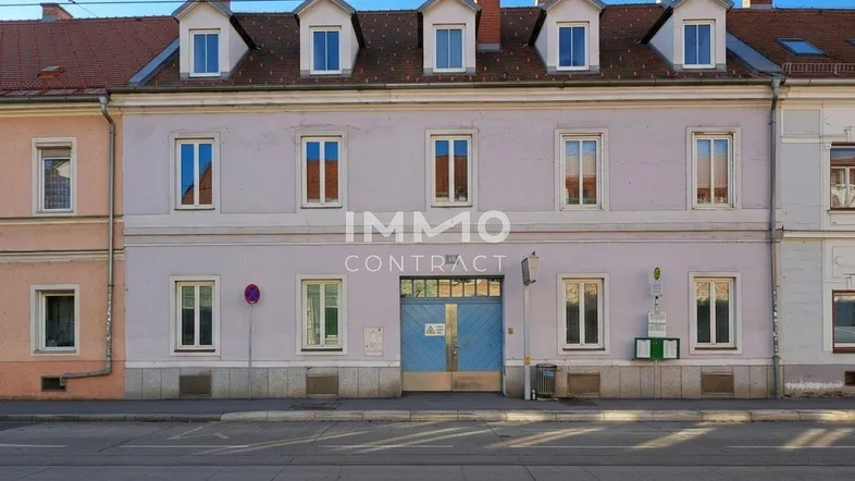 Fassade eines Altbaus mit mehreren Fenstern, Dachgauben und einem blauen Garagentor an der Straße.