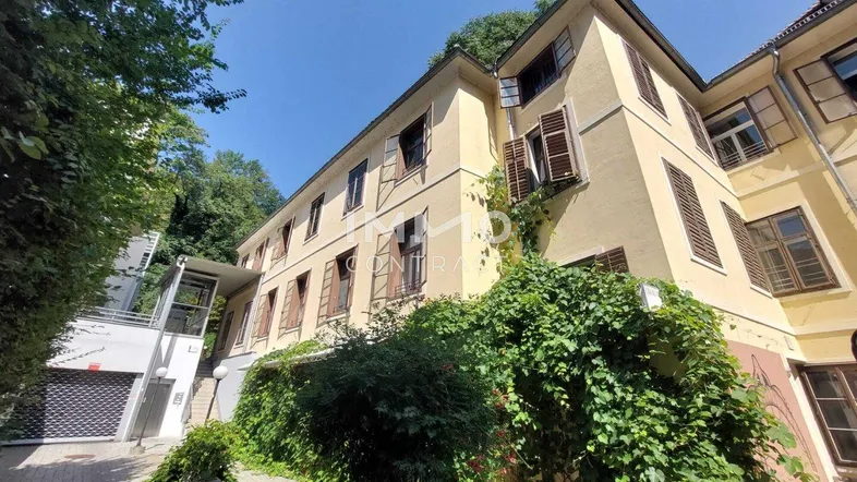 Gelbe Fassade eines Mehrfamilienhauses mit grüner Bepflanzung und einem Aufzug an der Seite.