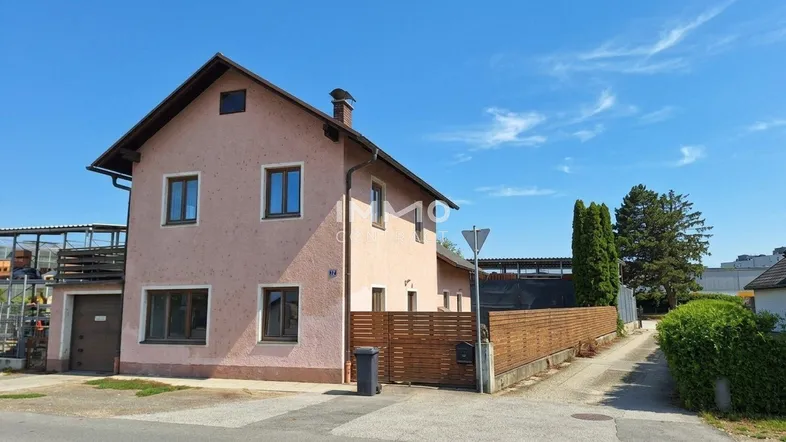 Vorderansicht eines Einfamilienhauses mit rosafarbener Fassade und Satteldach unter blauem Himmel.