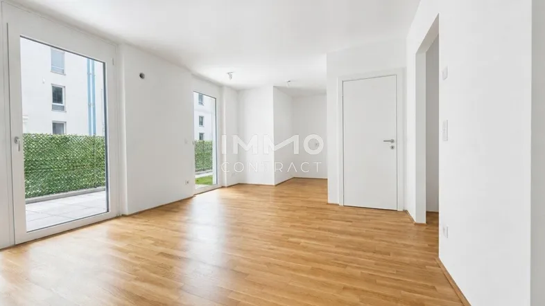Helles, leeres Zimmer mit Parkettboden und Zugang zu einer kleinen Terrasse mit Hecke.