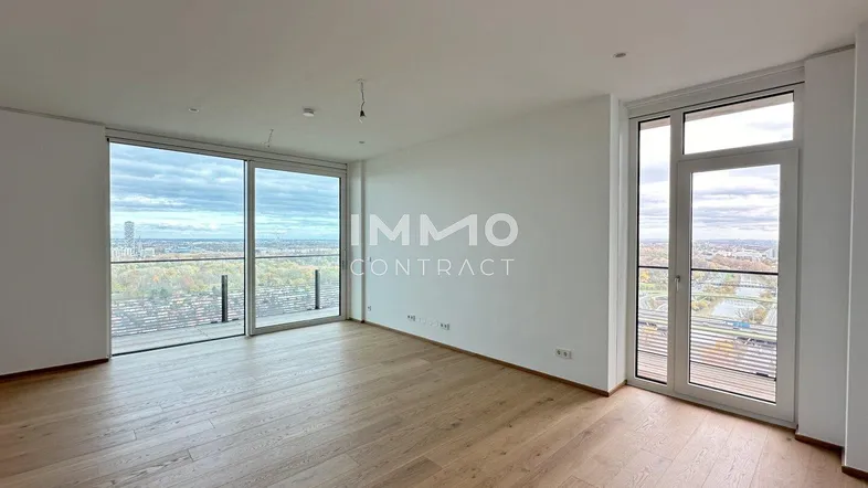 Helles, leeres Wohnzimmer mit großen Fenstern, Holzboden und weitem Blick über die Stadt und Grünflächen.