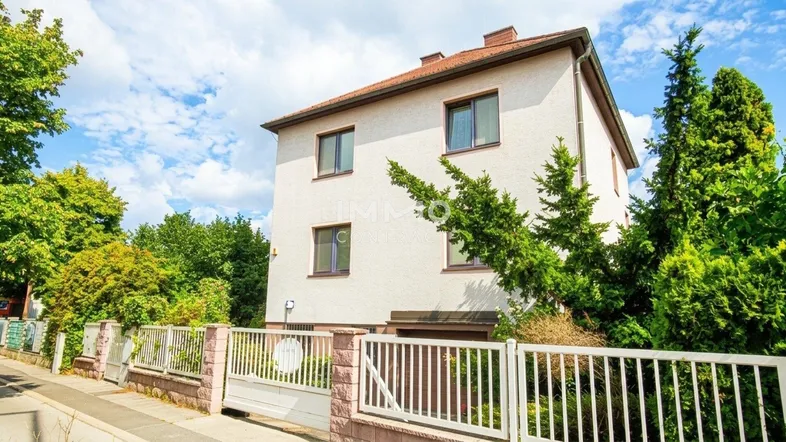 Weißes Einfamilienhaus mit braunem Dach, umgeben von einem gepflegten Garten und Zaun.