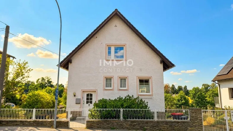 Gepflegtes weißes Einfamilienhaus mit Satteldach, kleinem Vorgarten und Zaun unter blauem Himmel.