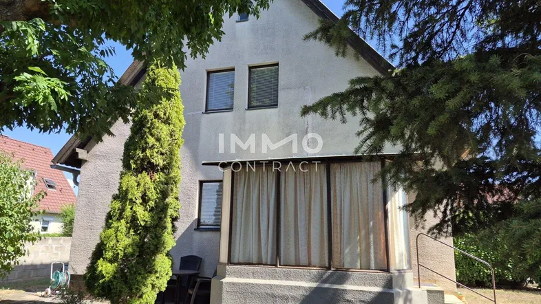 Seitenansicht des Hauses mit Fenstern, umgeben von Bäumen und einem kleinen Gartenbereich.