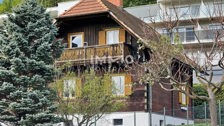 Traditionelles Holzhaus im Chalet-Stil mit Balkon, Ziegeldach und grüner Umgebung, im Hintergrund ein modernes Gebäude.