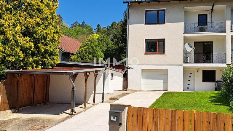 Mehrfamilienhaus mit weißer Fassade, Carport und gepflegtem Vorgarten unter blauem Himmel.