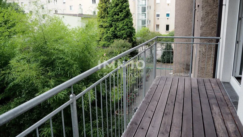 Balkon mit Holzboden und Metallgeländer, bietet einen grünen Ausblick und einen Aufzug im Hintergrund.