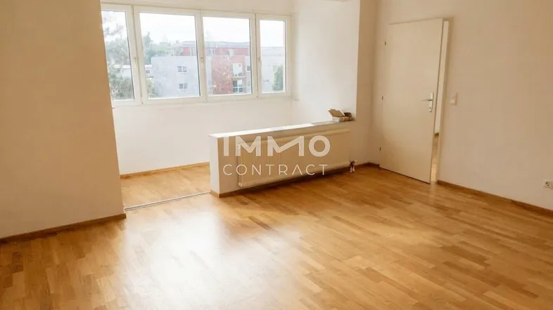 Helles, leeres Wohnzimmer mit Parkettboden und großen Fenstern, die viel Tageslicht hereinlassen.
