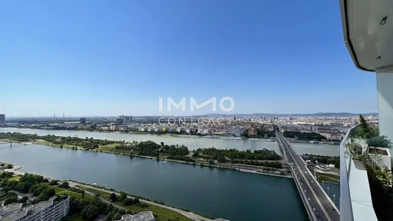 Panoramablick von einem Balkon auf die Donau, die Stadtlandschaft und eine Brücke unter blauem Himmel.