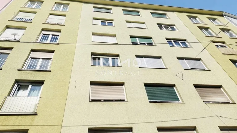 Fassade eines Mehrfamilienhauses mit zahlreichen Fenstern und Rollläden unter blauem Himmel.