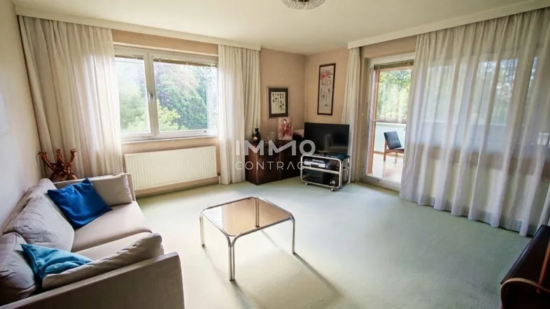 Helles Wohnzimmer mit Teppichboden, Sofa und Zugang zu einem Balkon mit Blick ins Grüne.
