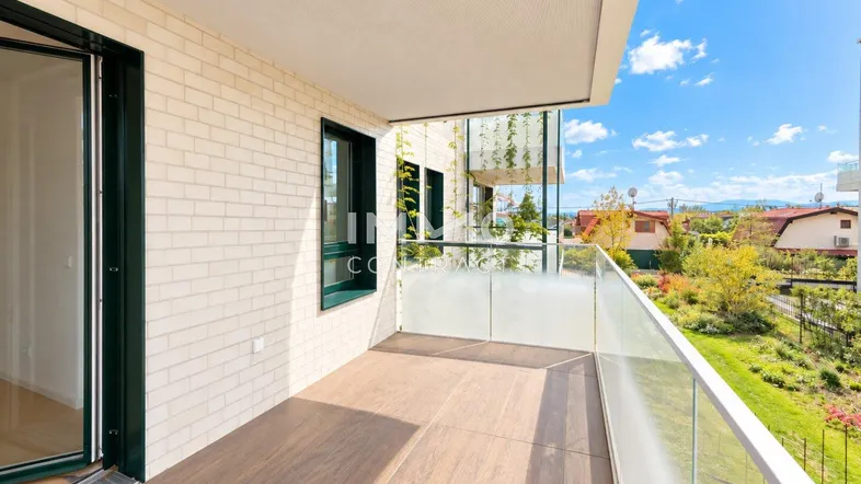 Heller Balkon mit Glasgeländer, Holzboden und weitem Blick ins Grüne.