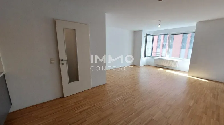 Helles, leeres Wohnzimmer mit Parkettboden und großen Fenstern, die viel Tageslicht hereinlassen.