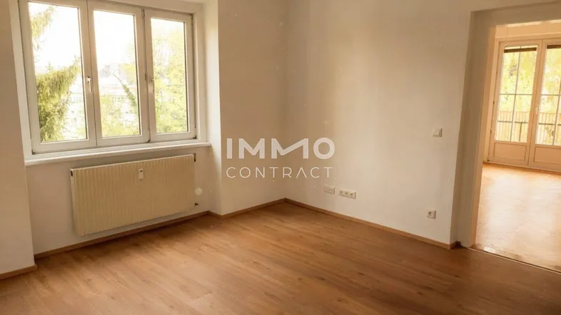Helles, leeres Zimmer mit Holzfußboden und großem Fenster, das viel Tageslicht hereinlässt.