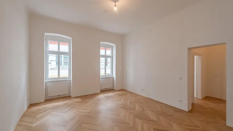 Helles, leeres Zimmer mit Fischgrätparkett und hohen Fenstern, bereit zum Einzug.