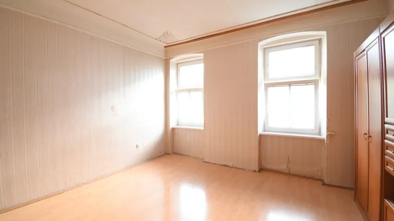 Helles Zimmer mit Laminatboden, zwei Fenstern und einer Stuckdecke, bereit zur Renovierung.
