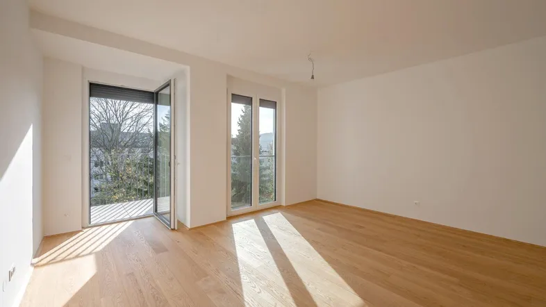 Helles Zimmer mit Holzboden und Zugang zu einem Balkon, durch große Fenster lichtdurchflutet.