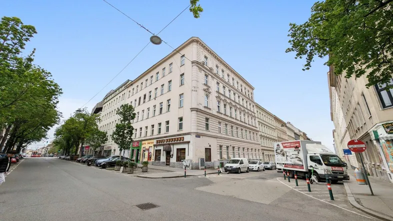 Historisches Eckgebäude an einer belebten Stadtstraße mit Bäumen und parkenden Autos.