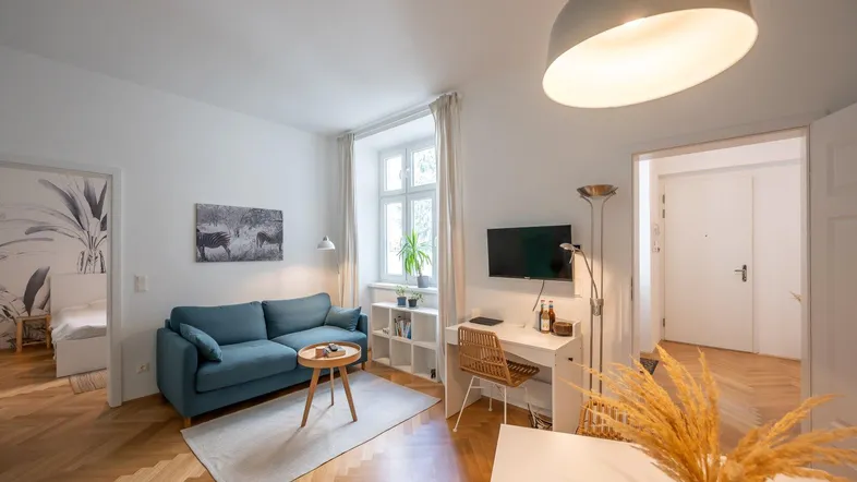 Helles Wohnzimmer mit blauem Sofa, Schreibtisch und Blick in ein angrenzendes Schlafzimmer.