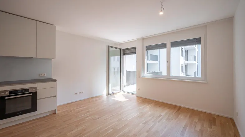 Heller Wohnbereich mit integrierter Küchenzeile, Parkettboden und Zugang zu einem Balkon.