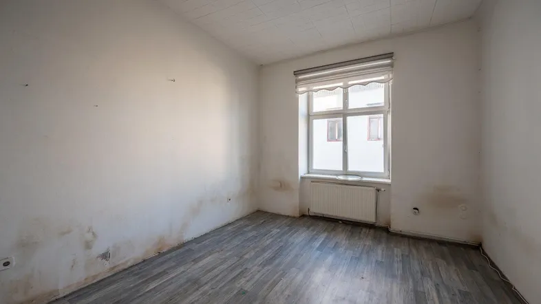 Leeres Zimmer mit Renovierungsbedarf, grauen Holzboden und einem Fenster mit Heizkörper.