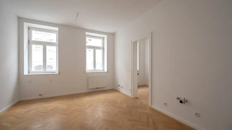 Helles Zimmer mit Fischgrätparkett, weißen Wänden und zwei Fenstern, bereit für die individuelle Gestaltung.
