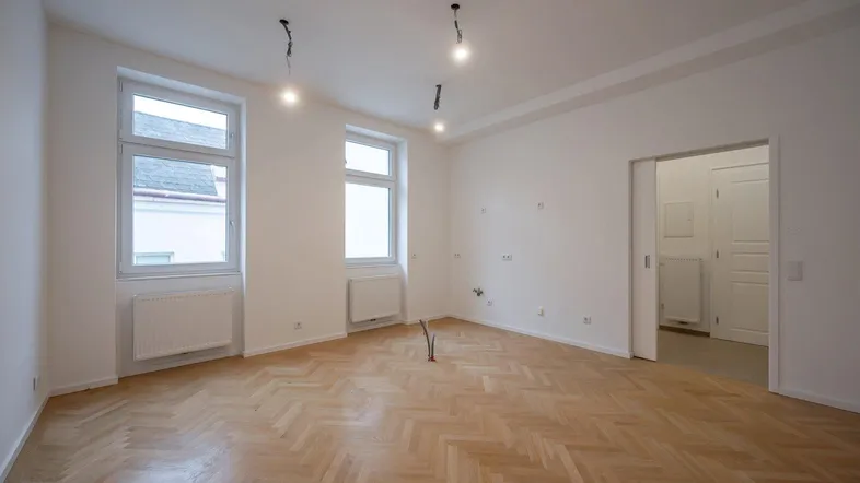 Helles, leeres Zimmer mit Fischgrätparkett und zwei großen Fenstern, bereit zur individuellen Gestaltung.