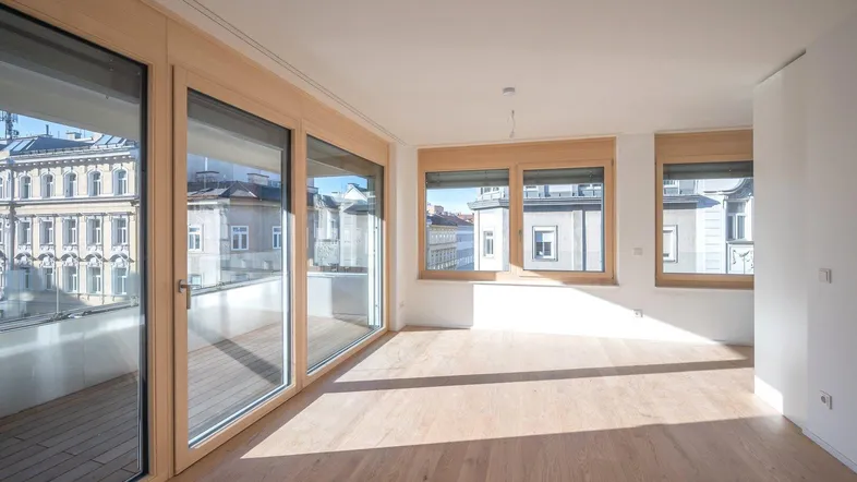 Helles, leeres Zimmer mit Holzboden und großen Fenstern, die viel Tageslicht hereinlassen.