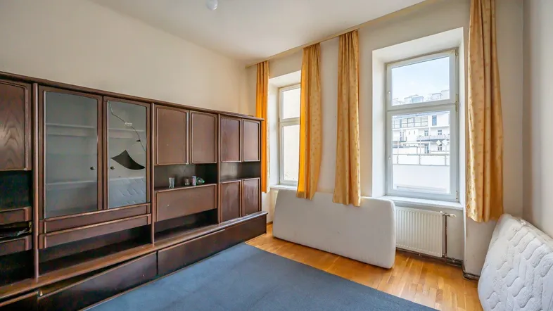 Geräumiges Wohnzimmer mit großem Schrank, Parkettboden und zwei hohen Fenstern.