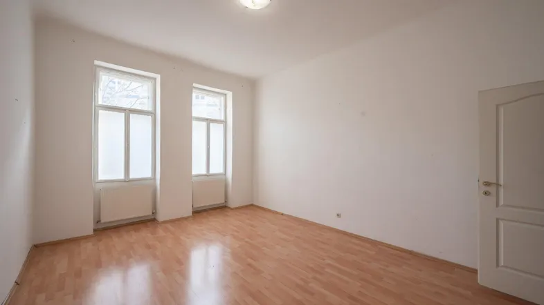Helles, leeres Zimmer mit Laminatboden und zwei Fenstern, bereit zur individuellen Gestaltung.