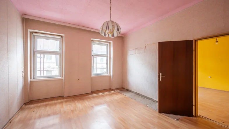 Leeres Zimmer mit Laminatboden, zwei Fenstern und rosa Decke, renovierungsbedürftig.