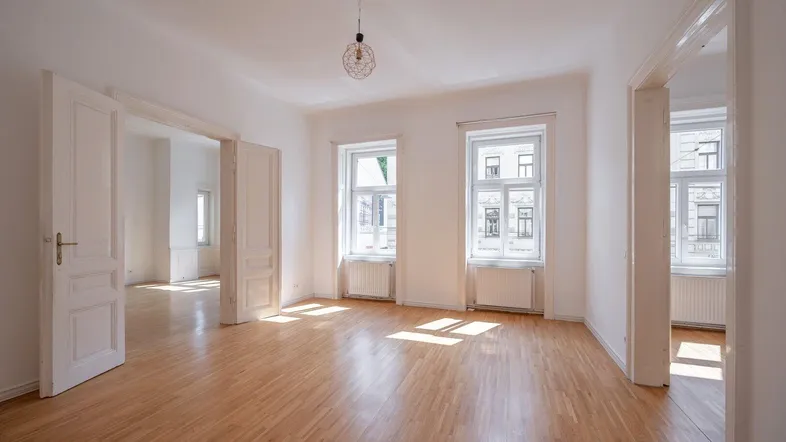 Helles, leeres Zimmer mit Parkettboden und hohen Fenstern, das viel Tageslicht hereinlässt.