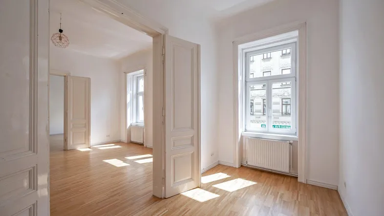 Helles, leeres Zimmer mit Parkettboden und hohen Fenstern, das viel Tageslicht hereinlässt.