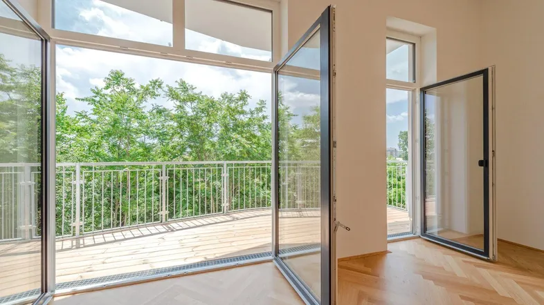 Helles Zimmer mit Fischgrätparkett und großen Fenstern, die auf einen grünen Balkon führen.