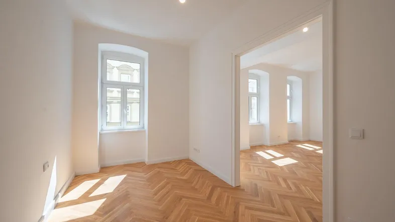 Helles, leeres Zimmer mit Fischgrätparkett und großen Fenstern, bereit zur individuellen Gestaltung.