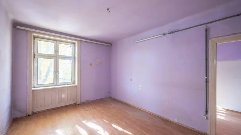 Leerer Raum mit lila Wänden, Holzboden und einem großen Fenster, das Tageslicht hereinlässt.