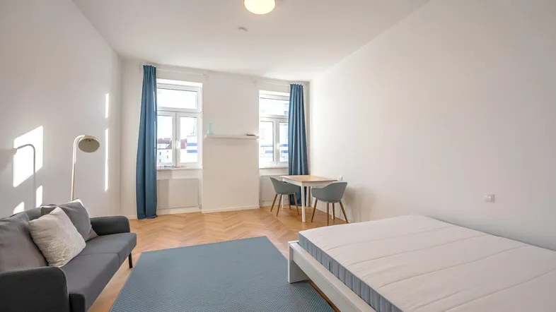 Helles Wohnzimmer mit Fischgrätparkett, Sofa, Bett und Esstischbereich, ideal für ein Studio-Apartment.