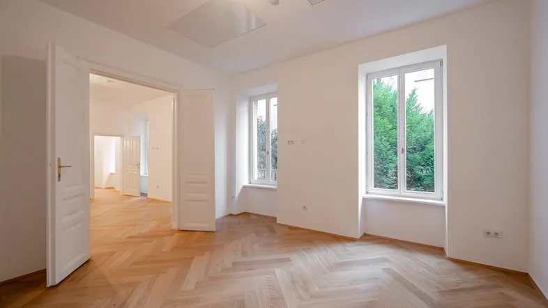Helles Zimmer mit Fischgrätparkett und hohen Fenstern, das viel Tageslicht hereinlässt.