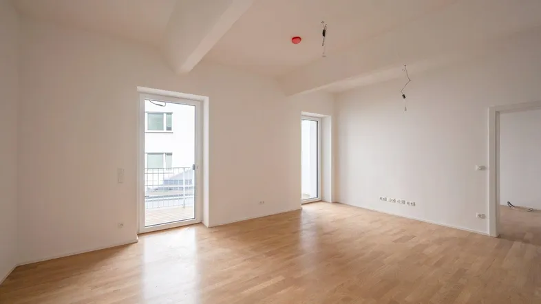 Helles, leeres Wohnzimmer mit Parkettboden und Zugang zu einem französischen Balkon.