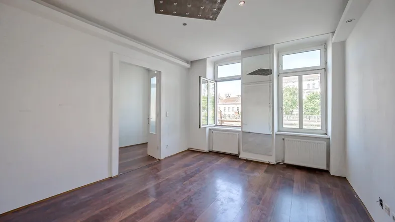 Helles, leeres Zimmer mit dunklem Holzboden, hohen Decken und Blick nach draußen.