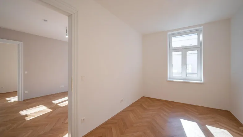 Helles, leeres Zimmer mit Fischgrätparkett und einem Fenster, das Tageslicht hereinlässt.