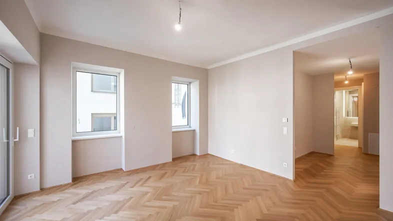 Helles, leeres Zimmer mit Fischgrätparkettboden und Blick in den Flur und ein Badezimmer.