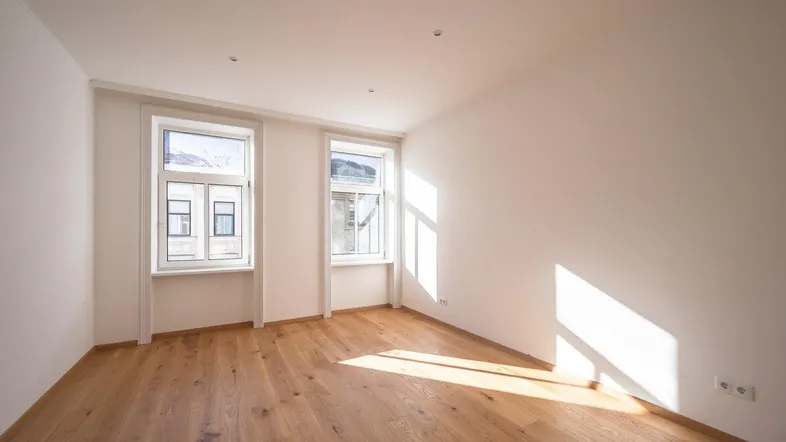 Helles, leeres Zimmer mit Holzboden und zwei Fenstern, durch die Sonnenlicht einfällt.