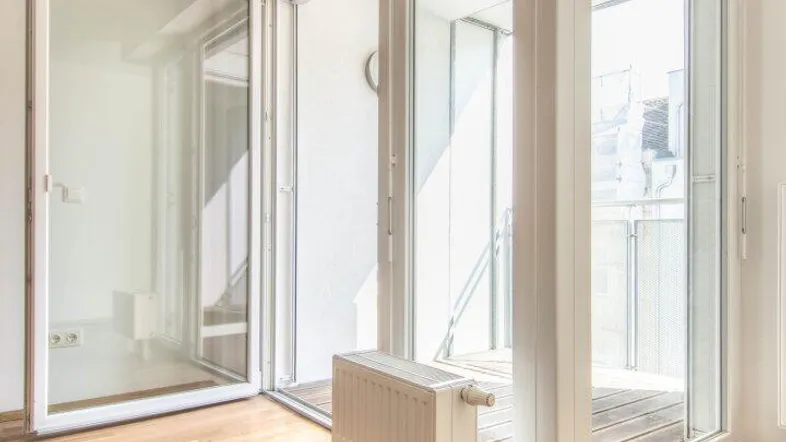 Helles Zimmer mit Parkettboden und Zugang zu einem Balkon, der viel Tageslicht hereinlässt.