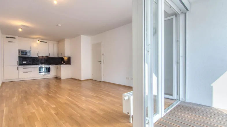 Heller offener Wohnbereich mit integrierter Küche, Parkettboden und Zugang zu einem Balkon.